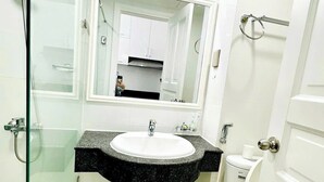 A bathroom with a glass shower enclosure, a white sink with a black countertop, a large mirror, and a white toilet. - Căn Hộ View Biển (Phú Thủy)