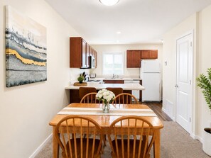 A compact kitchen with wooden cabinets, a white refrigerator, and a dining area with a table and chairs. - Holiday Village Townhomes – 3889 (Traverse City)