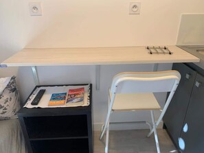Ein kleines, gemütliches Zimmer mit einem Schreibtisch, einem weißen Stuhl, einem schwarzen Schrank und einer Fernbedienung auf einem kleinen Tisch.