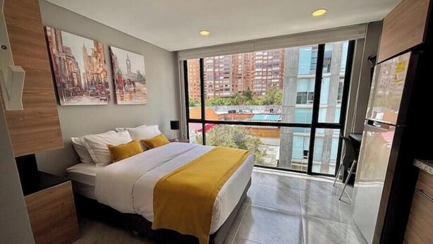 Un dormitorio con cama, mesita de noche, ventana con vista a la ciudad y refrigerador.