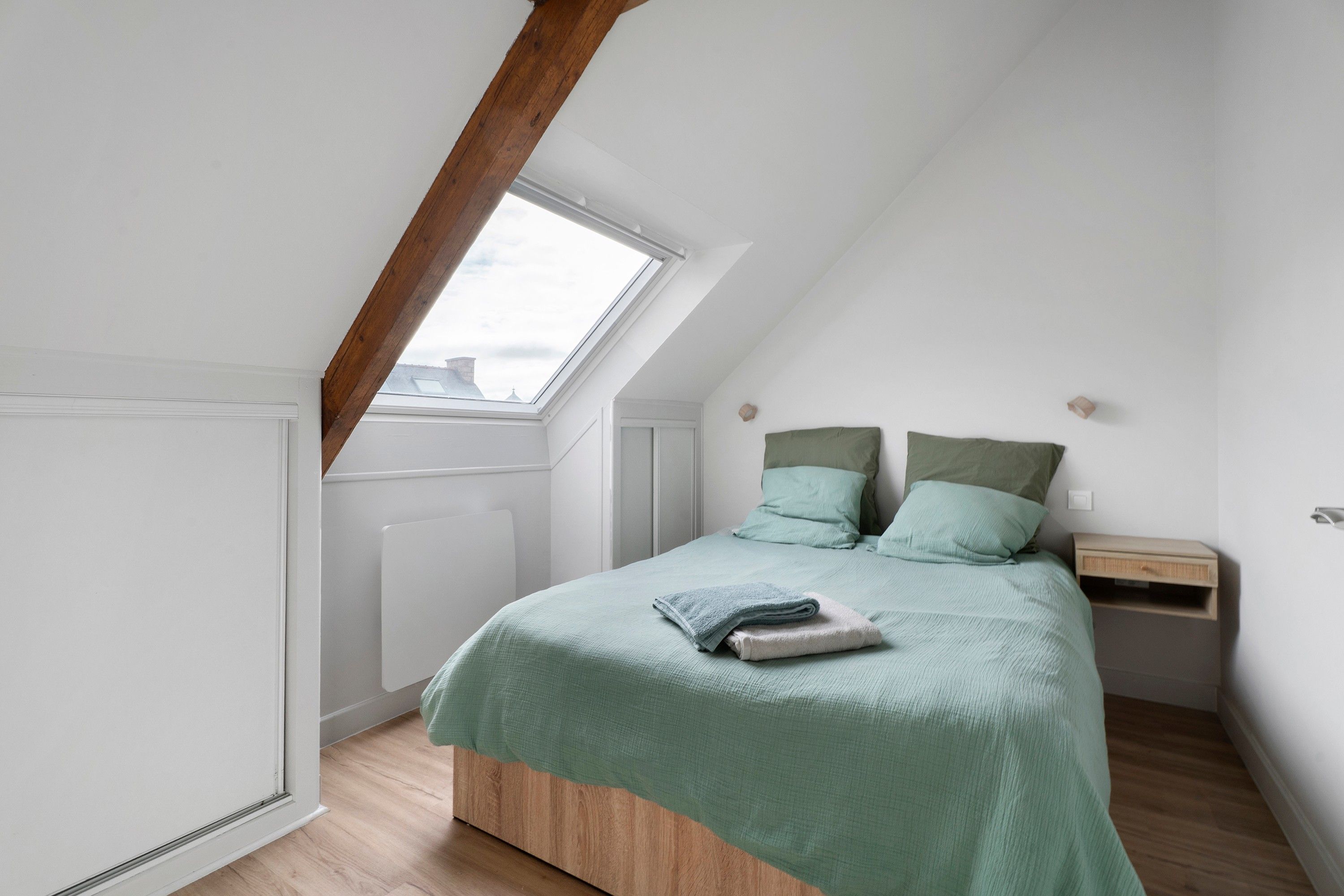 A bedroom with a sloped ceiling, a skylight, a bed with green bedding, a nightstand, and a wooden floor.