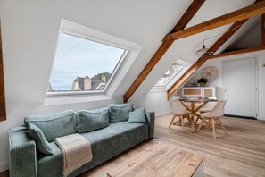 Ein Loft-Zimmer mit schrägem Dach, einem grünen Sofa, einem Esstisch mit Stühlen und Blick auf Gebäude durch ein Dachfenster.