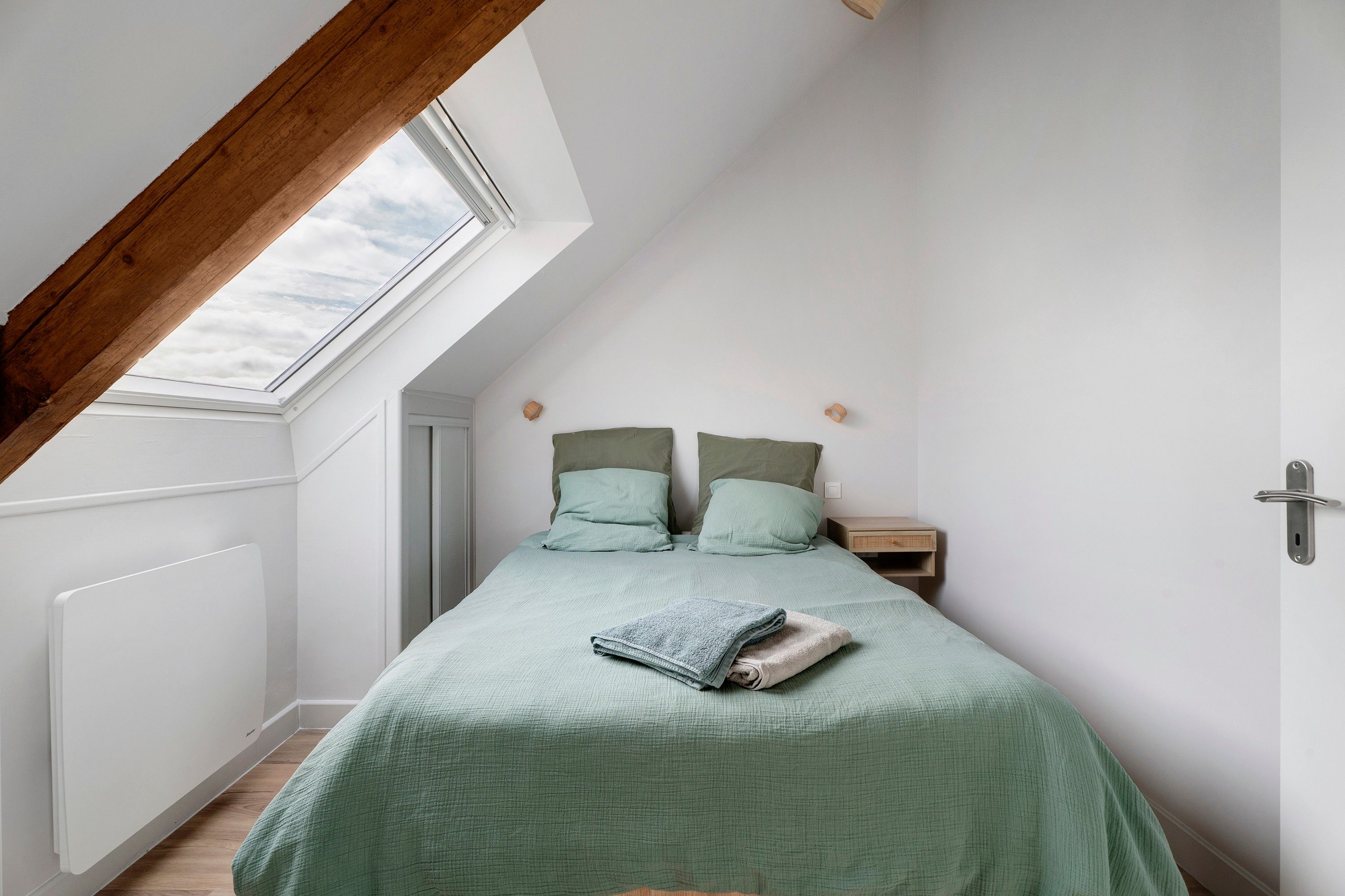 Un dormitorio con techo inclinado, tragaluz, cama con ropa de cama verde, mesita de noche y piso de madera.