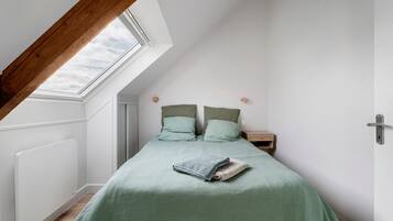 Un dormitorio con techo inclinado, tragaluz, cama con ropa de cama verde, mesita de noche y piso de madera.