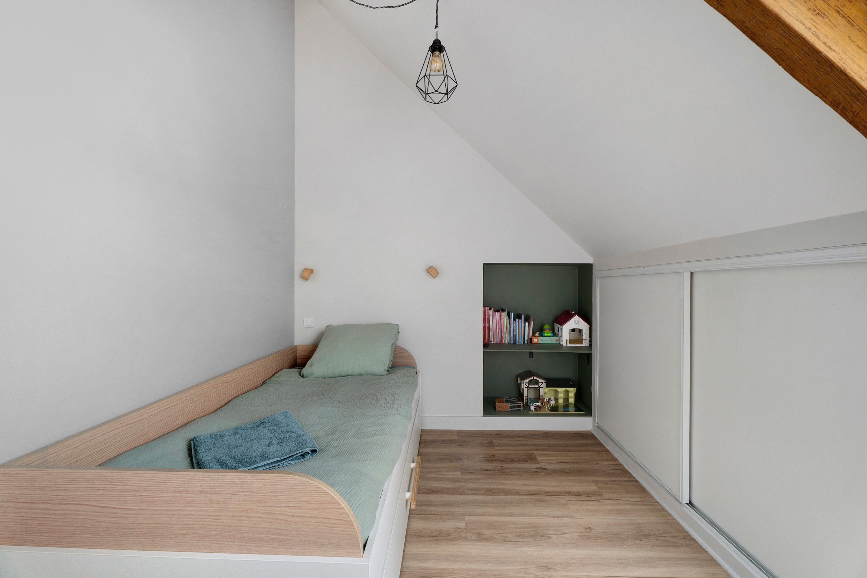 Une petite chambre à coucher accueillante, avec un plafond en pente, un lit simple, une petite bibliothèque et une fenêtre.