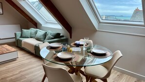A dining area with a glass table, white chairs, and a view of the sea through skylights. - Apartment 'Les Fougères' with Sea View and Wi-Fi (Erquy)