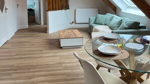A loft-style room with a sloped ceiling, wooden beams, a glass dining table, white chairs, a sofa with cushions, a TV mounted on the wall, and a skylight. - Apartment 'Les Fougères' with Sea View and Wi-Fi (Erquy)