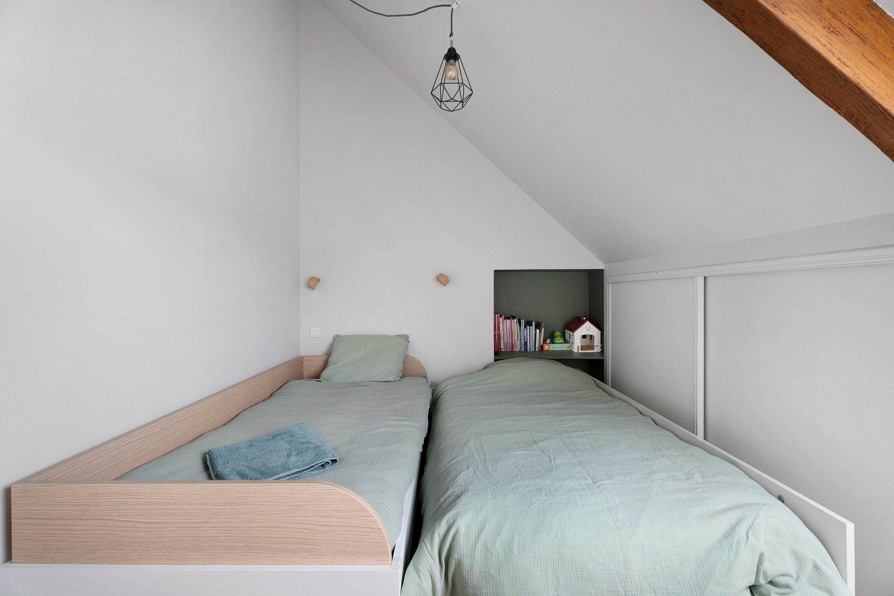 A small, cozy bedroom with a sloped ceiling, a single bed, a bedside table, and a wall-mounted shelf with books and a small house model.