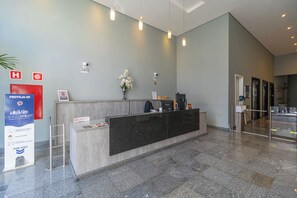 Un comptoir d’accueil avec une pancarte indiquant « PROTEJA-SE » et « LIVRAISON », dans un hall d’entrée moderne avec un mur gris, un vase de fleurs et une caméra de surveillance.