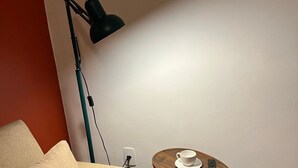 Ein Zimmer mit einem beigefarbenen Sofa, einem kleinen runden Tisch mit Tasse und Fernbedienung, einer Stehlampe und einem Holzboden.