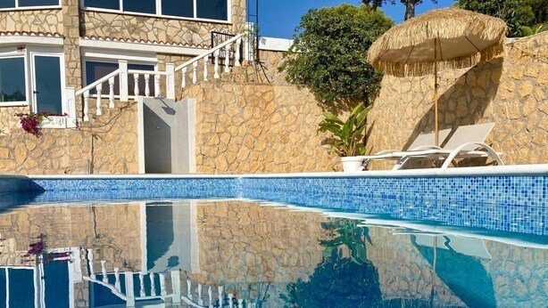 Una piscina con agua azul cristalina, paredes de piedra, una escalera blanca y un paraguas de paja.