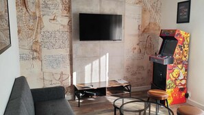 Sala de estar com TV de tela plana, console de jogos, sofá, mesa de centro e parede com desenhos técnicos.