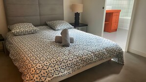 Ein ordentlich bezogenes Bett mit gemusterten Betttextilien, eine Nachttischlampe und Blick auf einen gefliesten Boden sowie eine Tür zu einem anderen Raum.
