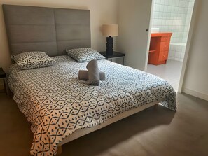 Una cama bien hecha con ropa de cama estampada, una lámpara de noche y vistas de un suelo de baldosas y una puerta que da a otra habitación.