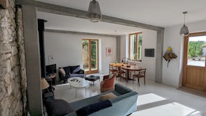 A spacious living area with a sofa, coffee table, and dining table. There are large windows providing natural light and a view of greenery outside. - La Maison des Remparts: Private Pool, Wi-Fi, Air Conditioning – Holiday Home in Clérieux (Clérieux)