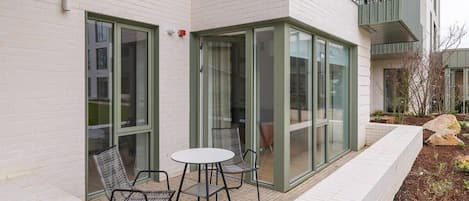 A modern building with a glass door entrance, a small patio with a table and chairs, and a brick pathway.