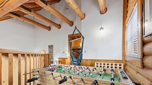 Una habitación con piso de madera, una mesa de fútbol de mesa, vigas de madera y un ventanal con persianas.