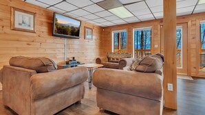 A log cabin interior with a TV mounted on the wall, leather sofas, a small table, and large windows offering views of trees. - A Family Affair #712 (Gatlinburg)