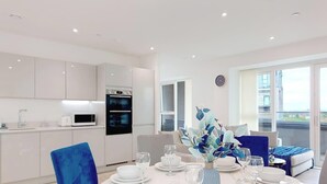 A modern kitchen with white cabinets and a black oven, a dining area with a table set for four, and a balcony with a view of the city. - Deluxe Serviced Three Bedroom Apartment (London)