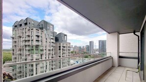 A balcony with a view of high-rise buildings, a glass railing, and a chair. - Deluxe Serviced Three Bedroom Apartment (London)