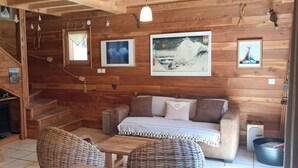 Interno di una baita in legno con divano, poltrone in vimini, camino e quadri appesi alle pareti.