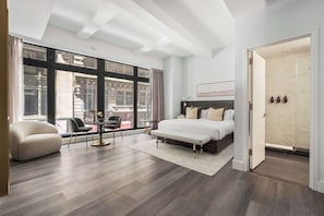 Ein modernes Schlafzimmer mit einem großen Bett, einer Couch, einem Sessel und Blick auf die Stadt durch große Fenster.