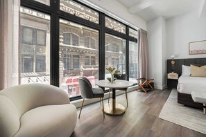 Ein modernes Hotelzimmer mit großem Fenster, einem runden Esstisch, einer Couch, einem Bett und einem Nachttisch.