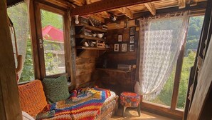 Interior acogedor de una cabaña con techo de madera, un banco empotrado con una manta colorida, estantes con libros y objetos decorativos, y una vista a través de un ventanal grande.