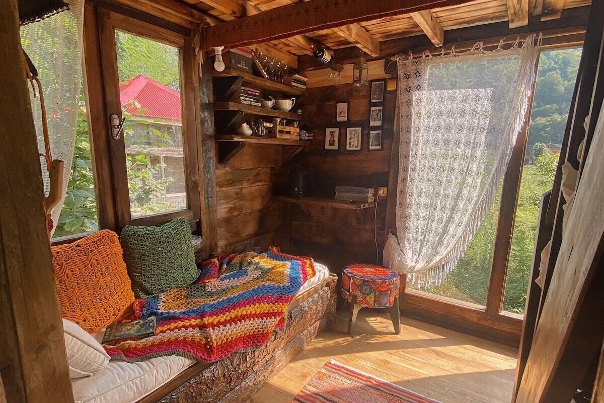 A cozy cabin interior with a wooden ceiling, a built-in bench with a colorful blanket, shelves with books and decorative items, and a view through a large window.