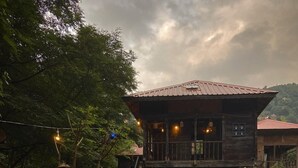 Una casa de madera rústica con un balcón, rodeada de follaje y una silla de madera.