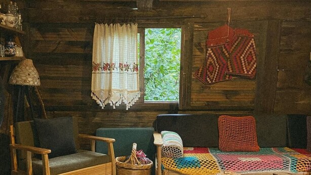 Interior rústico de una cabaña con paredes de madera, un sofá verde, una silla de madera, una cesta tejida y una alfombra estampada.