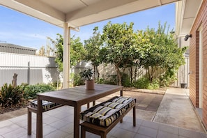 Eine überdachte Terrasse mit Holztisch und gepolsterten Bänken, umgeben von einem Garten und einer Ziegelmauer.