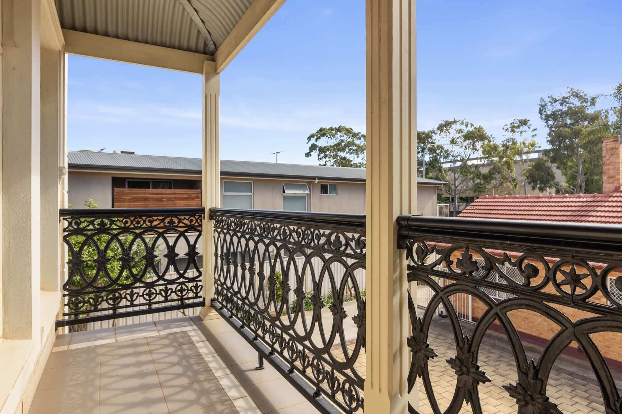 A covered balcony with ornate black metal railings, a tiled floor, and a view of residential buildings in the background.