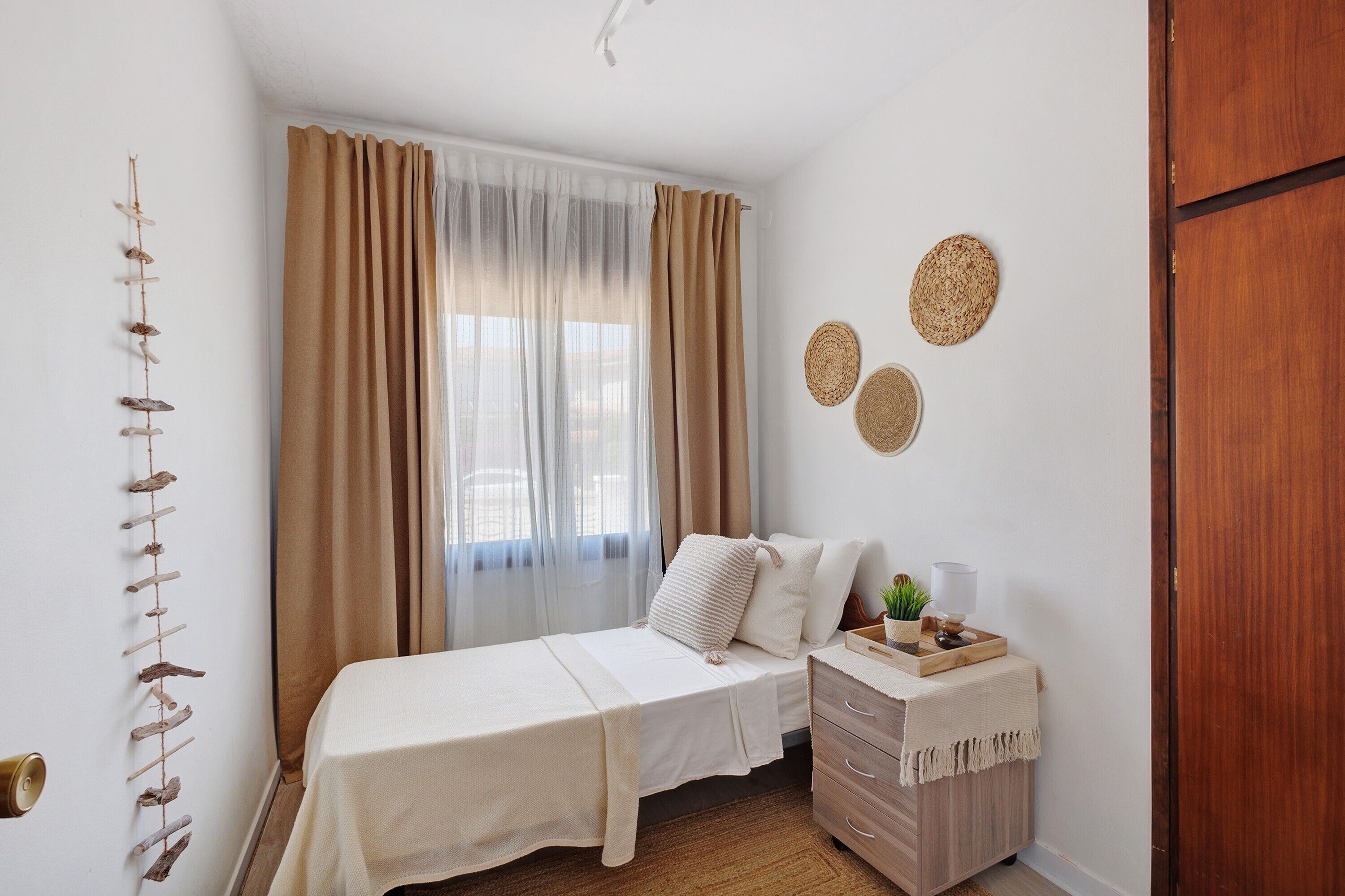 Un dormitorio con cama, mesita de noche, planta en la pared y ventana con cortinas.