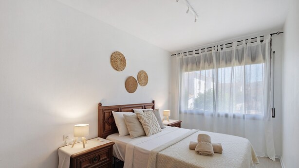 A bedroom with a wooden bed, bedside tables with lamps, a window with sheer curtains, and wall decor.