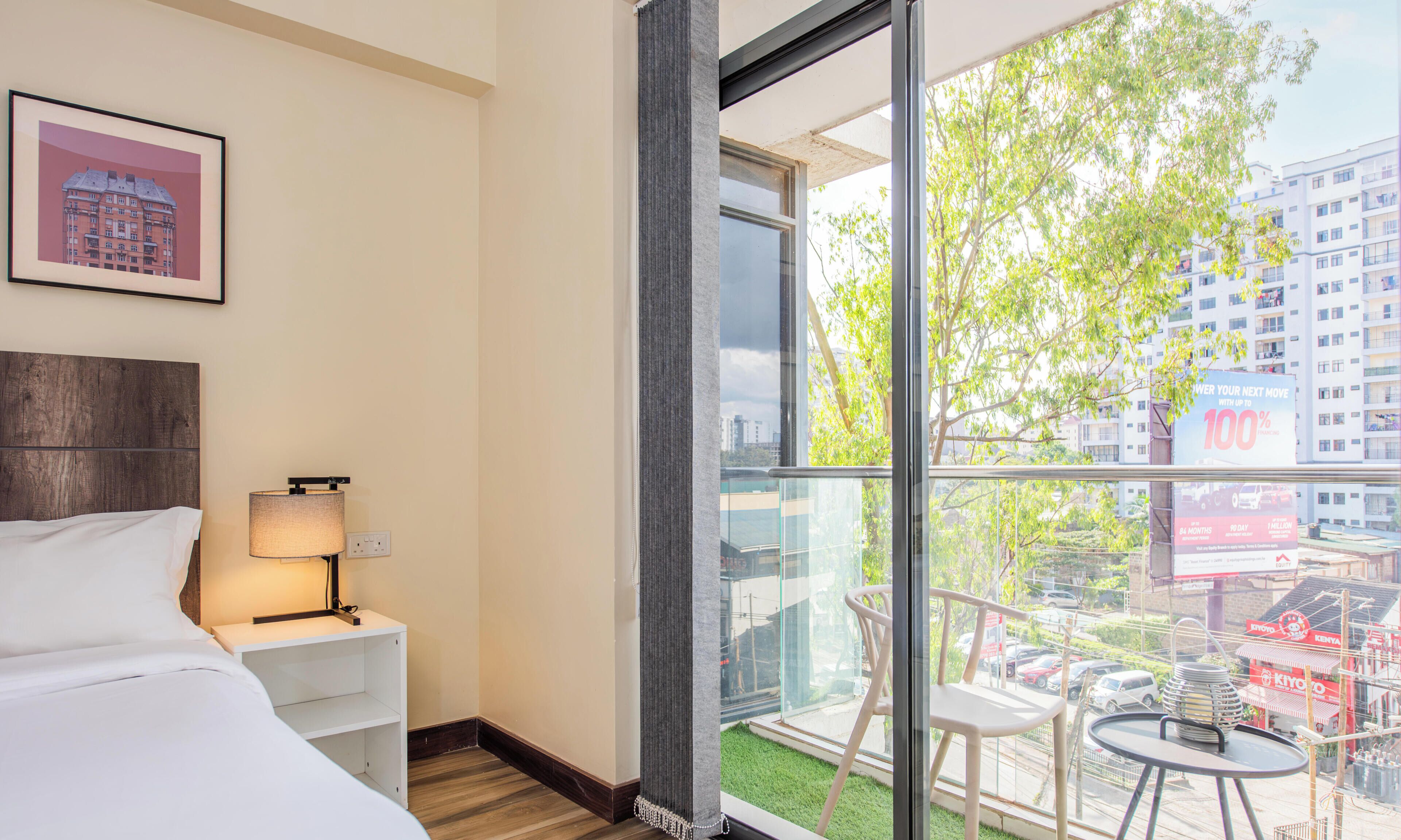 Hotelkamer met een bed, een bedlampje, een ingelijst schilderij aan de muur en een groot raam met uitzicht op de stad en groen.