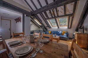 Ein Essbereich mit Holztisch, gedeckt für vier Personen, eine Küche im Hintergrund und ein Schlafzimmer im oberen Bereich mit einer Couch und Blick ins Freie.