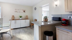 Une cuisine moderne avec des meubles blancs, une poubelle en acier inoxydable, une machine à café rouge et un coin repas avec une table et des chaises en verre.
