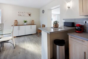 A modern kitchen with white cabinets, a stainless steel trash bin, a red coffee maker, and a dining area with a glass table and chairs.