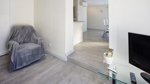 Un salon moderne avec un fauteuil recouvert de tissu gris, une table basse en verre avec des télécommandes, et un téléviseur à écran plat fixé au mur.