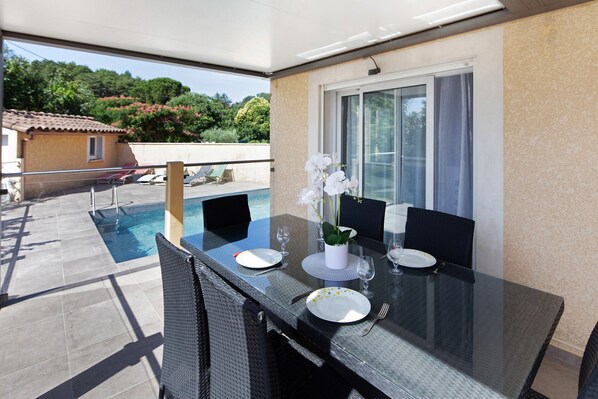 A glass-top dining table with chairs, a pool, and a view of a house and trees.