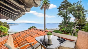 Eine Terrasse mit geflastertem Dach, einem Tisch mit Topfpflanze und Blick auf das Meer und Bäume.