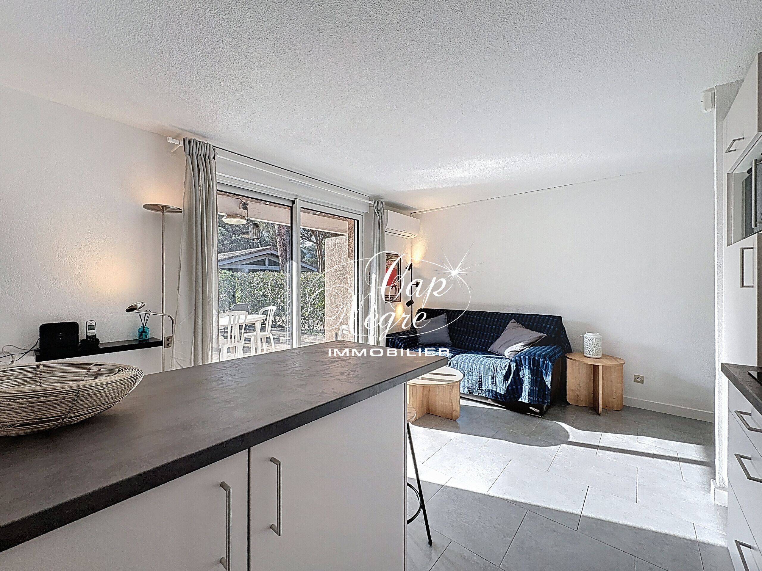 Une cuisine moderne dotée d’un grand îlot central, de portes-fenêtres coulissantes donnant sur une terrasse et d’un coin salon confortable avec un canapé et des tables d’appoint.