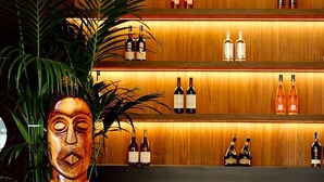 A bar area with wooden shelves displaying bottles, two bar stools, and a wooden sculpture. - MBH | Laguna King Suite (Diso Spongano)