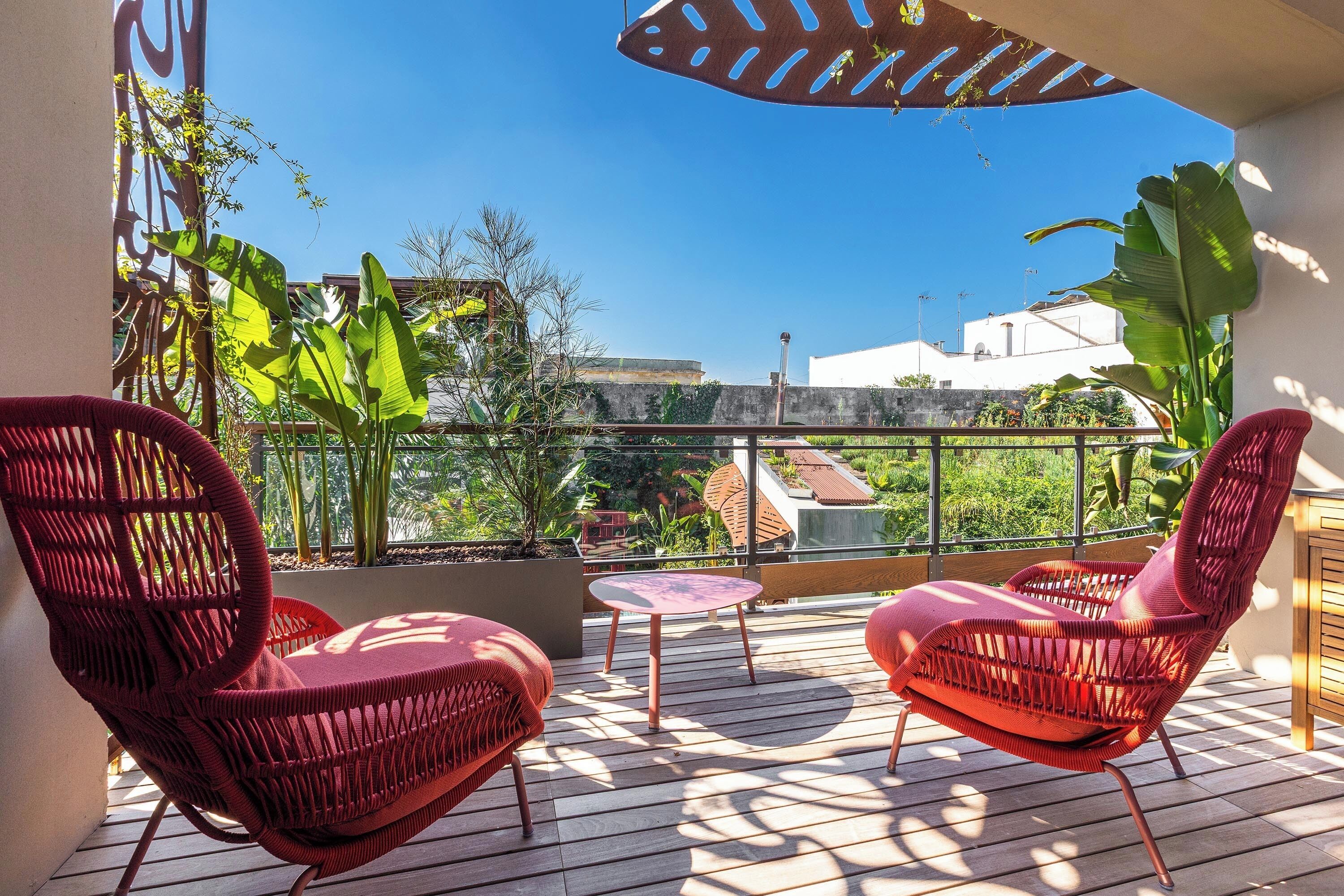 A balcony with two red wicker chairs, a small round table, and a view of buildings and greenery.