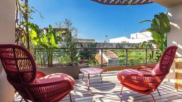 A balcony with two red wicker chairs, a small round table, and a view of buildings and greenery.