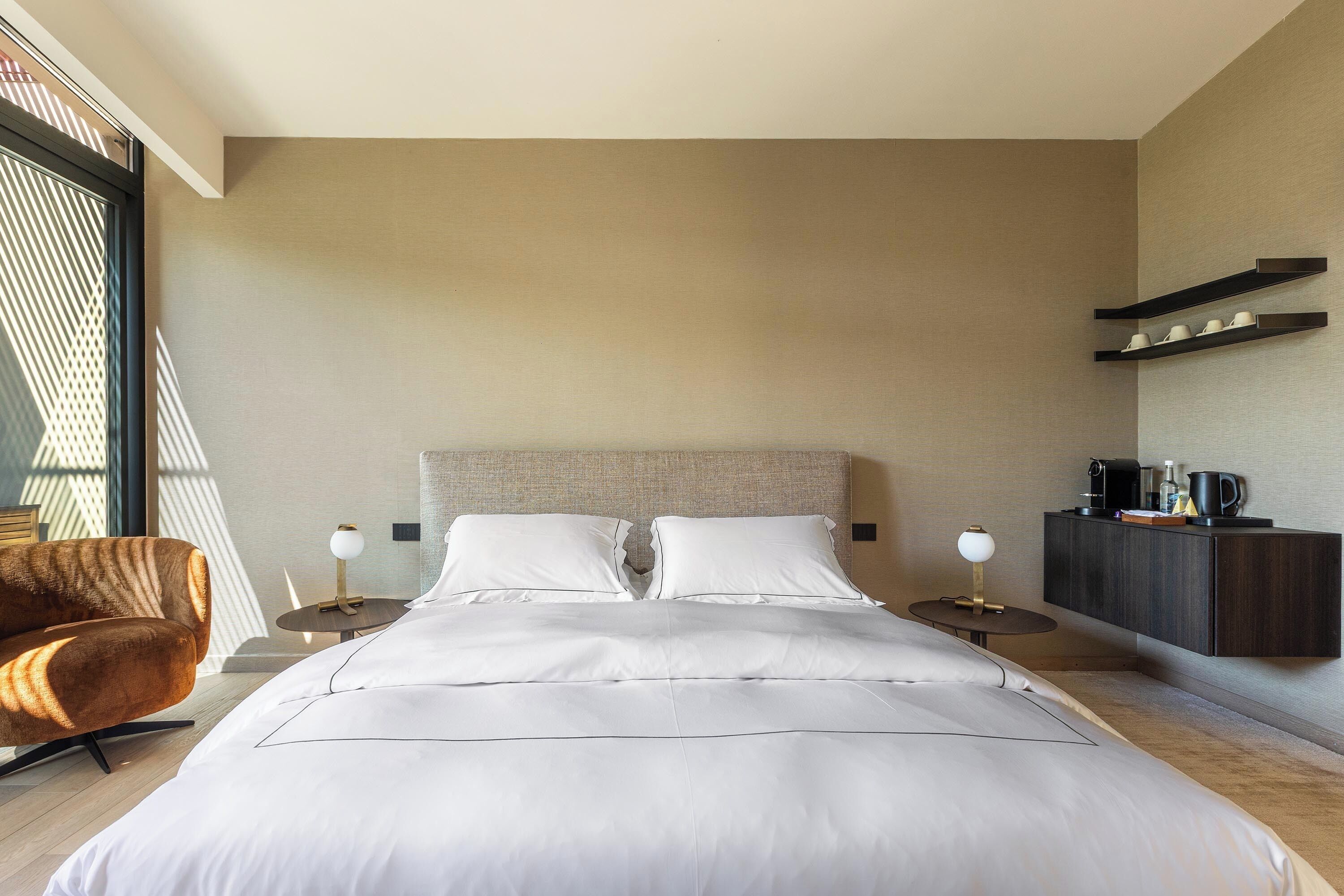 Ein modernes Hotelzimmer mit einem großen Bett, zwei Nachttischen, einem Sessel und einem Kleiderschrank, mit Blick durch ein Fenster.