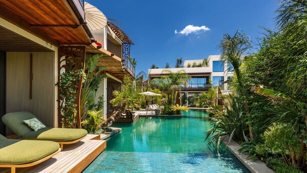 A pool with lounge chairs, surrounded by greenery and a building with balconies.