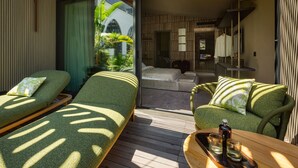 A room with two green cushioned beds, a wooden table with glasses, and a view of a balcony with plants.