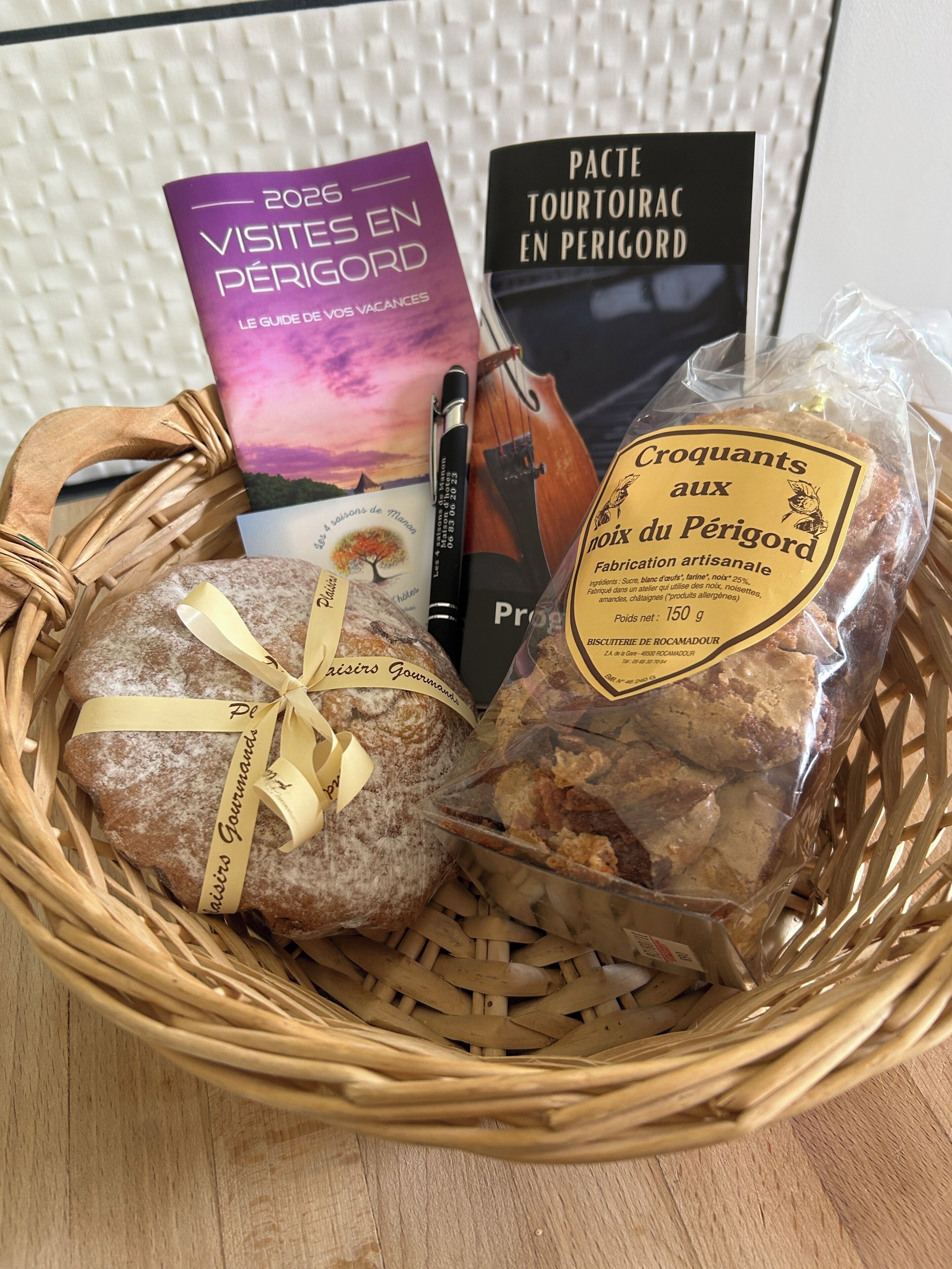 A wicker basket containing a loaf of bread, a book titled 'Pacte Tourtoirac en Perigord,' and a package of croissants.